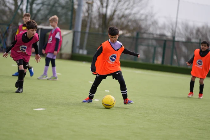 A boy in an orange foundation bib dribbles with the ball, as players in pink bibs close him down