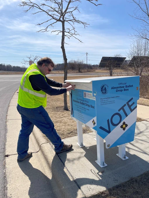 Election official in a yellow safety vest locking a secure official absentee ballot drop box outdoors on a sunny day.