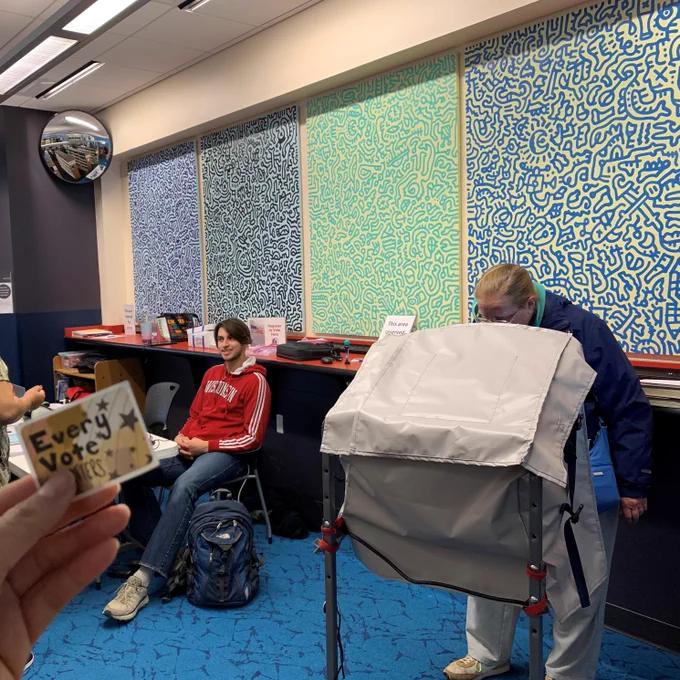 Voter at an accessible ballot marking device marking their ballot inside the Goodman South Library room with colorful abstract wall art and a seated poll worker nearby.
