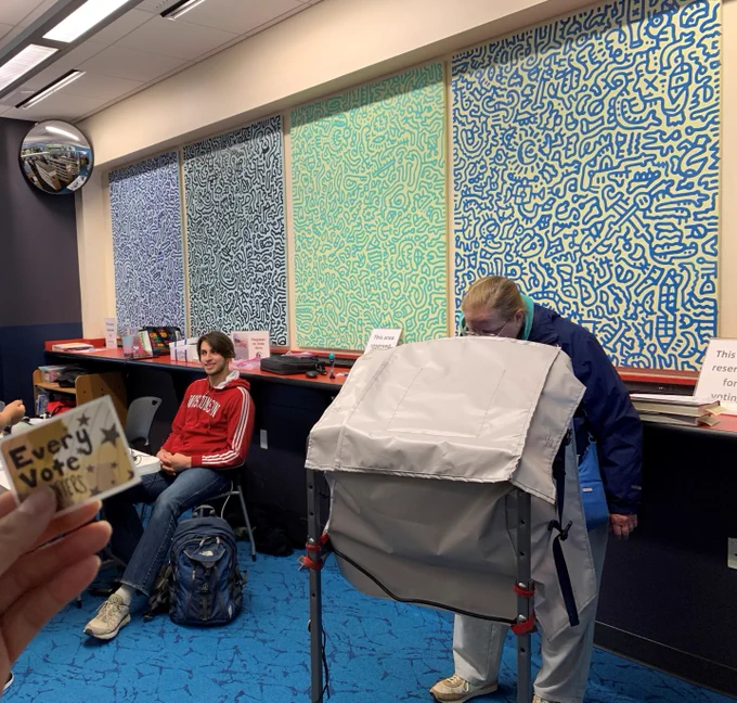 Voter at an accessible ballot marking device marking their ballot inside the Goodman South Library room with colorful abstract wall art and a seated poll worker nearby.