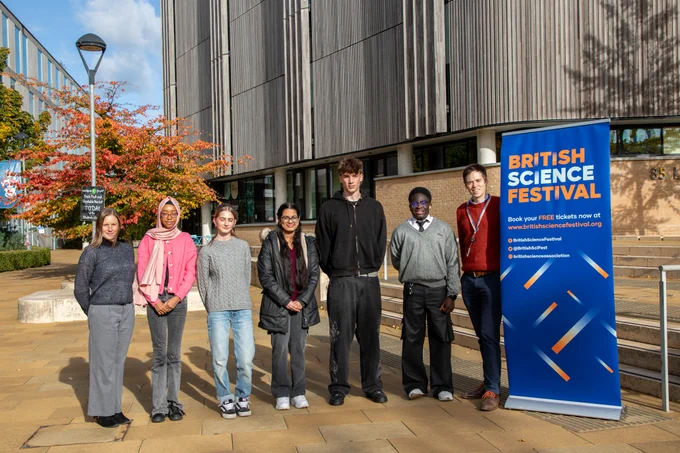 A group of seven people stands in front of a modern building beside a blue banner reading "British Science Festival." They are outdoors under a blue sky with a vibrant tree showing autumn colors behind them.
