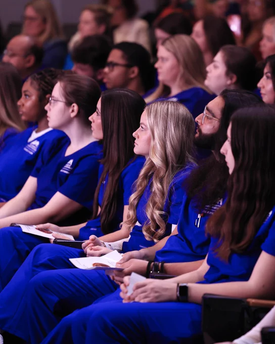 A  group of nursing students in blue scrubs attentively listening during the Spring 2026 White Coat Ceremony. 