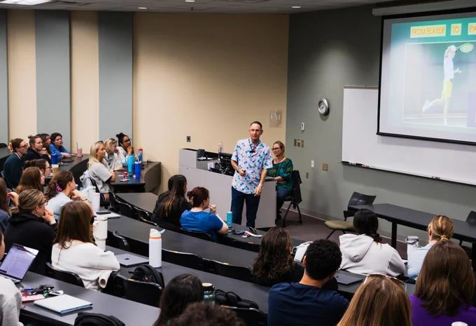 A classroom with a speaker standing in front of seated attendees. A presentation slide shows an image of a tennis scene. Attendees appear engaged, with some using laptops and taking notes.