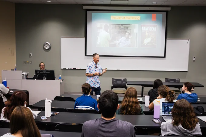 A person stands at the front of a classroom giving a presentation. The presentation slide shows images from the movie "Raw Deal." Students sit at desks listening, and the class instructor is seated at a computer.