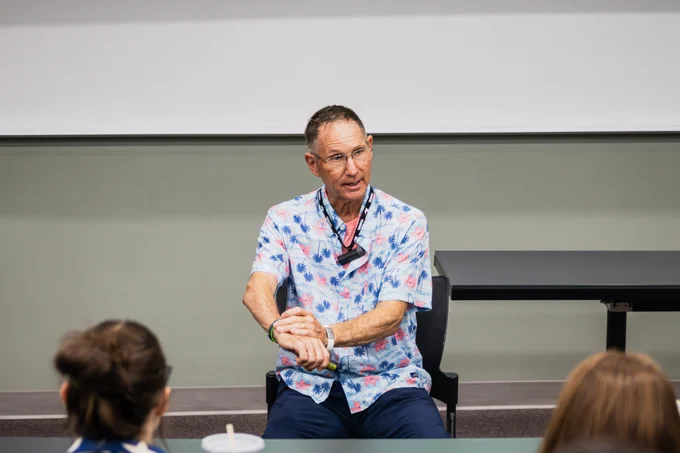 A person in a colorful patterned shirt sits at the front of a classroom, speaking to an audience. A couple of individuals are seated in the foreground, listening. The setting is a lecture room with a table and a cup visible.