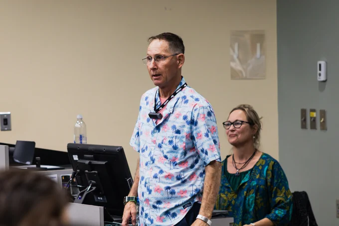 A person in a tropical shirt stands at a podium in a classroom, speaking. Another person with glasses and a green shirt sits nearby, smiling. Computers and classroom equipment are visible in the background.