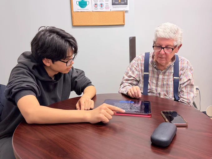 Photo of a teenage boy (left) helping an older adult (right) use an iPad.