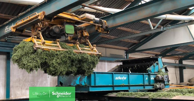Ein Kran bef\u00F6rdert Gras in einen blauen Container in einer Halle. 