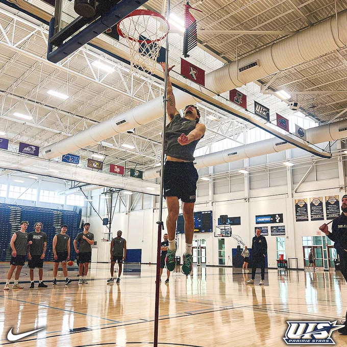 A basketball player jumps to touch a vertical measuring device near the hoop in an indoor gym, while teammates and coaches watch from the court behind him.