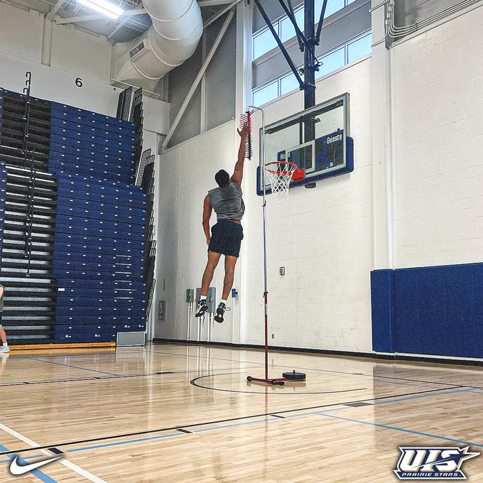 A basketball player jumps to touch a vertical measuring device near the hoop in an indoor gym, while teammates and coaches watch from the court behind him.