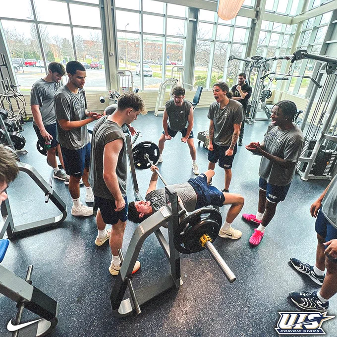 A group of basketball players surrounds a teammate performing a bench press in a bright weight room, cheering him on as he lifts the bar.