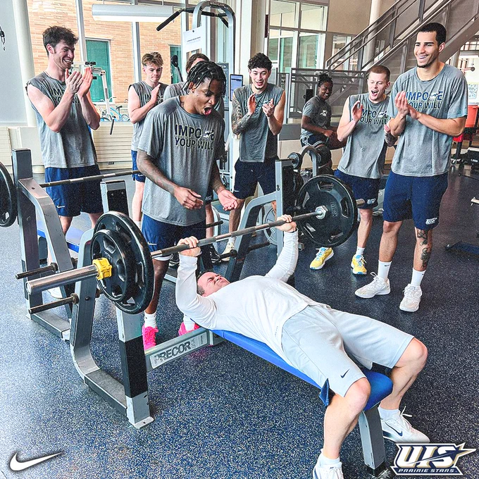 A group of basketball players surrounds a teammate performing a bench press in a bright weight room, cheering him on as he lifts the bar.