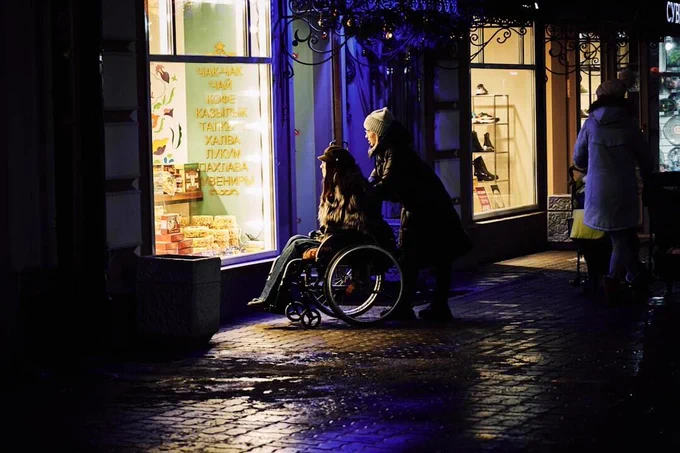 A person pushes another person in a wheelchair along a wet, illuminated street at night. They pause in front of a brightly lit shop window displaying items and glowing yellow signage.