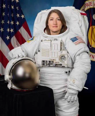 Official portrait of astronaut Christina Koch, dressed in a white spacesuit with her hand on top of a helmet. The American flag is seen in the background. 