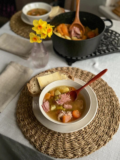 Welsh ham cawl (a gammon ham and root vegetable broth) in a bowl, with a big pan of cawl and some daffodils in a vase just behind it.