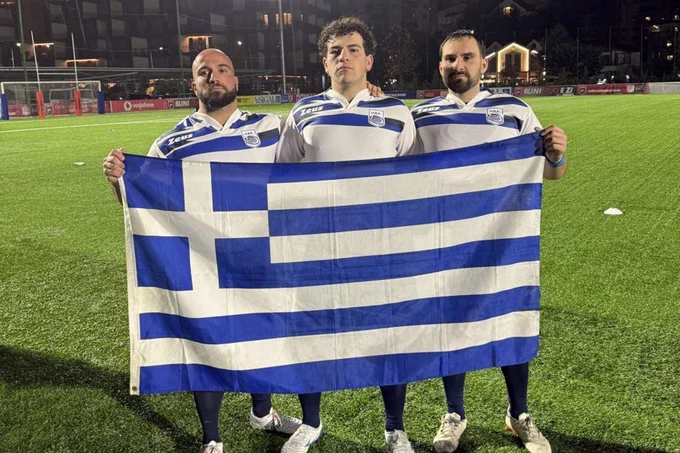 Three men hold up a Greek flag while standing on a sports field. 