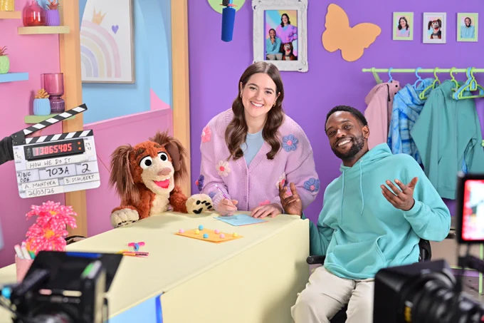 Donovan, Allison and Howie the Dog pose on set of How We Do It, a pink and purple space filled with colourful accessories.