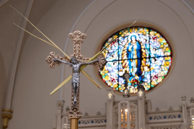 Photo inside the Chapel of the Most Holy Rosary with a silver crucifix adorned with palm fronds in the foreground against a blurred, colorful stained-glass window depicting a religious figure. 