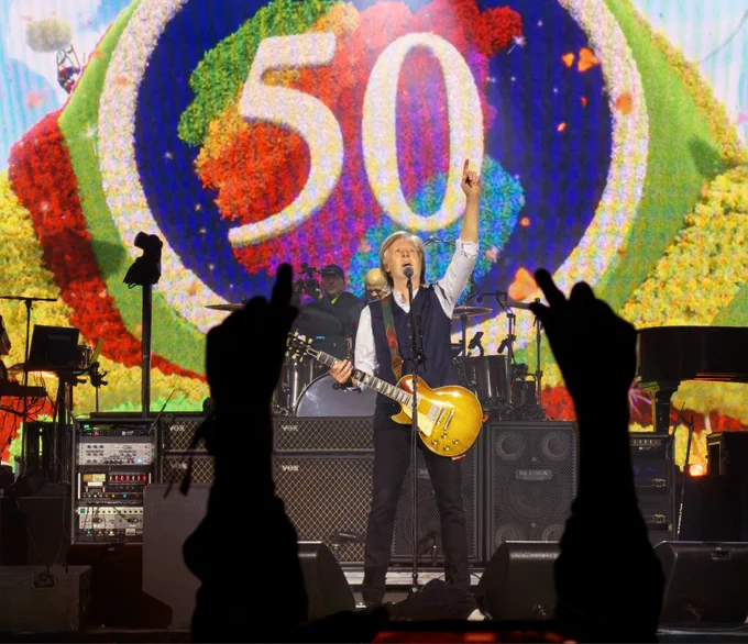 Sir Paul McCartney on stage at Apple Park, playing guitar and pointing to the sky, with a large “50” behind him embroidered on the Apple logo.

