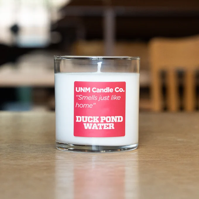 A glass candle labeled "UNM Candle Co. 'Smells just like home' - Duck Pond Water" on a table.