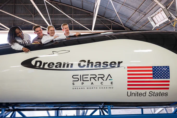 A group of people lean out of the window of the Sierra Space Dream Chaser spacecraft. The side of the spacecraft features the American flag, the "Dream Chaser" name, and the Sierra Space logo.