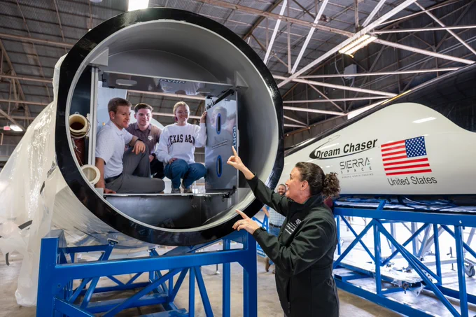 Workers examine the interior of a Dream Chaser spacecraft. Three individuals are seated inside a cylindrical part of the craft, while another person stands outside, pointing at a section of the structure. The spacecraft is in a large industrial setting, with visible support beams and equipment.