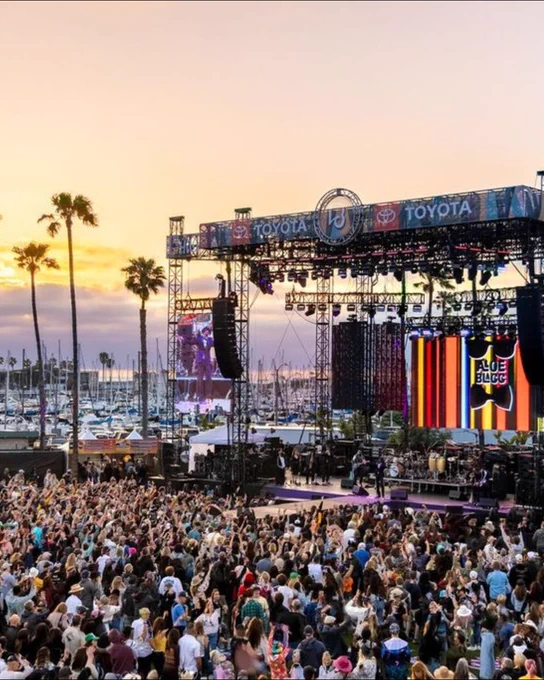 A large crowd gathers at an outdoor music festival during sunset. Palm trees and sailboats in a marina are visible in the background.
