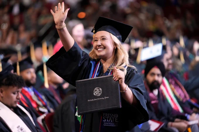 A person in a graduation cap and gown waves while holding a diploma from Fresno State. Other graduates are visible in the background, all seated in an auditorium.