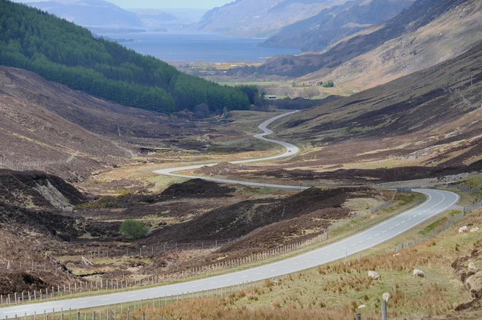 Glen Docherty. The image shows a view down the glen with the road sweeping broadly from side to side. There’s part of a forest on the left in the middle distance and a large loch flanked by mountains at the top of the frame.