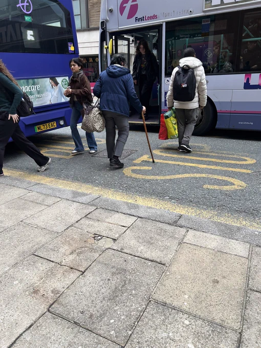 Passengers getting on and off the bus in the middle of the road. A passenger walking in the road toward the bus is using a walking stick.