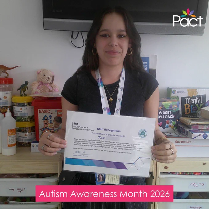 A woman stands indoors holding a printed staff recognition certificate. She is wearing a lanyard and jewellery, and is positioned in front of shelves filled with toys, art supplies, and children’s books. The Pact logo appears in the top right corner, and a pink banner at the bottom reads “Autism Awareness Month 2026.”