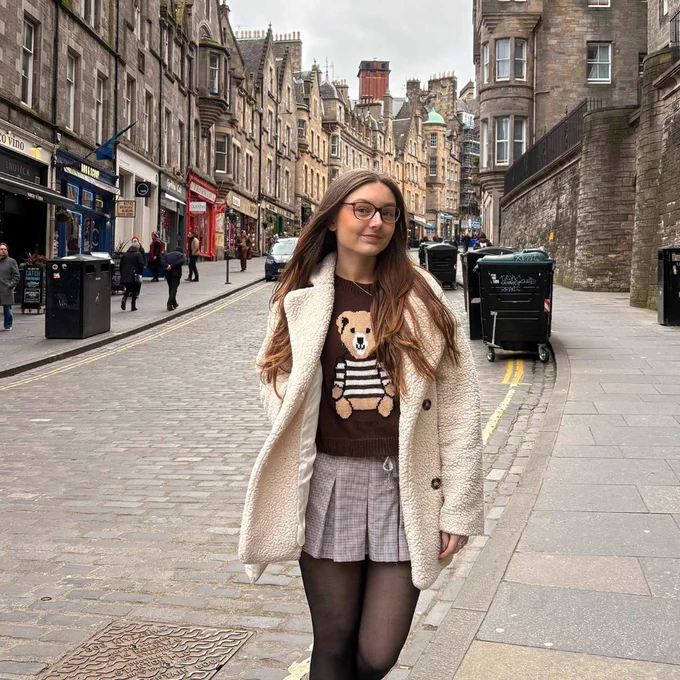 Ulster University student Bethany wearing a teddy bear sweater, beige coat and grey skirt walking on a cobblestone street lined with historic buildings and shops.