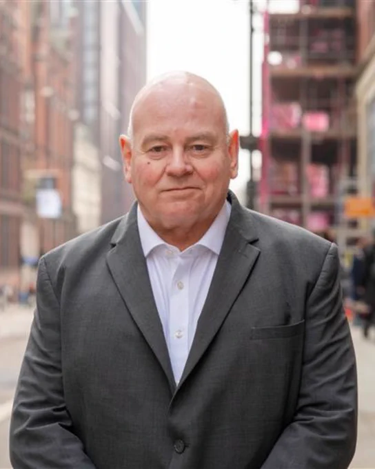 A person in a dark suit stands on a city street, facing the camera with a neutral expression.