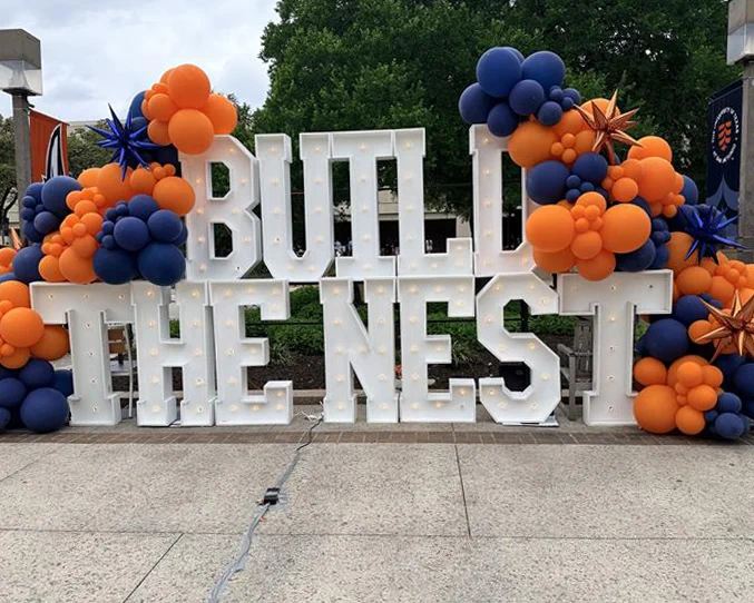 A large installation with lit-up letters spells "BUILD THE NEST," surrounded by clusters of blue and orange balloons and star-shaped decorations.