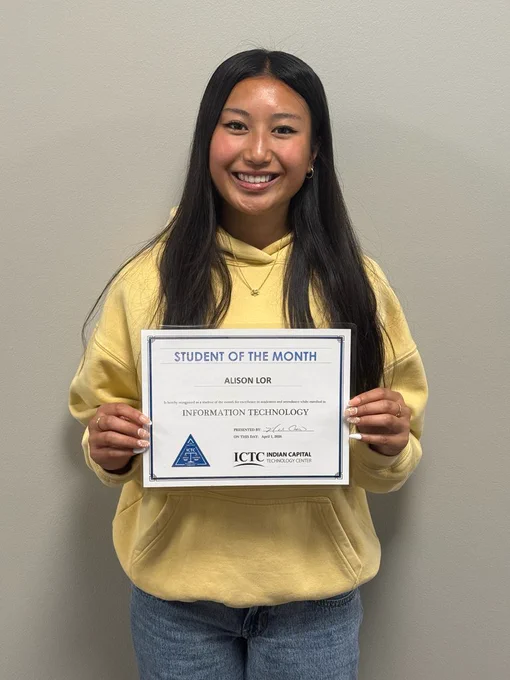 A person with long hair holds a certificate for student of the month. They are smiling and wearing a yellow sweater.