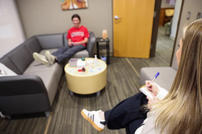 A therapist sits on a chair, writing notes while observing a college student across the room on another couch.