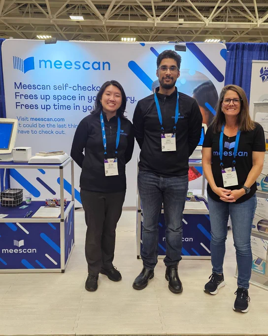 Three Meescan team members stand smiling at their booth during a conference. They are positioned in front of a large branded backdrop that reads “meescan” and highlights self-checkout benefits. Each person is wearing a name badge and dark Meescan-branded clothing. On either side of them are display tables with self-checkout devices and materials. The setting appears to be a convention center exhibit hall with overhead lighting and booth structures visible in the background.