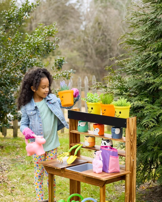 Child wearing pink gloves tending to smiling yellow plant pots on a wooden outdoor gardening table surrounded by greenery.