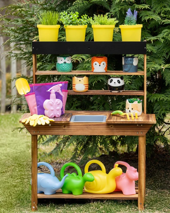 Wooden gardening station with yellow pots of herbs, animal-themed planters, colorful watering cans, and gardening tools arranged outdoors.
