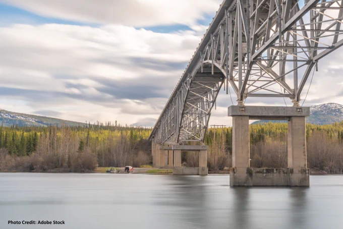 A large steel bridge spans across a calm river, supported by multiple concrete pillars. In the background, a forest of green trees lines the riverbank, and snow-capped mountains are visible under a cloudy sky.