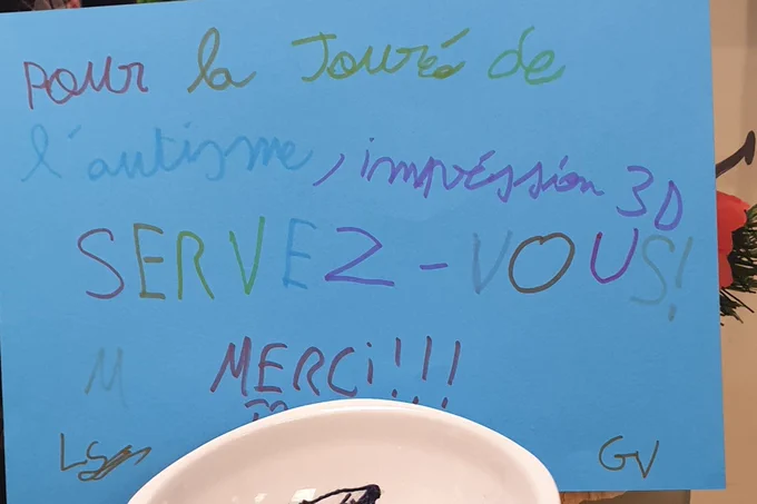 Affiche faite à la main sur une feuille bleue avec des messages colorés en soutien à l’autisme, dont « Pour la journée de l’autisme, impression 3d, servez-vous ! » et « Merci ». En bas, un bol blanc contenant des bracelets bleus. 