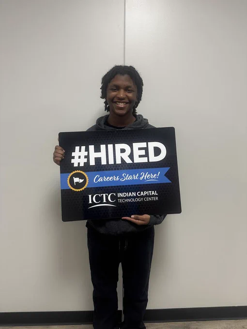 Person with dark hoodie holds sign that says "#HIRED Careers Start Here!" and logo. White background.