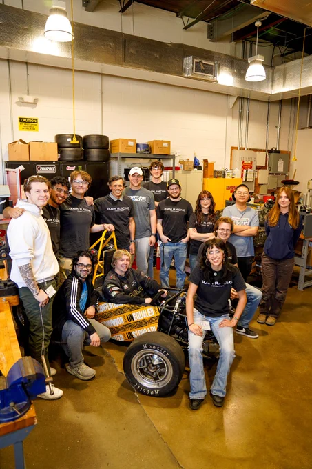 Dr. Rao and a large group of students pose around a black and gold V-C-U branded Formula One race car. One student sits in the car. Several students wear black Ram Racing shirts and safety goggles.