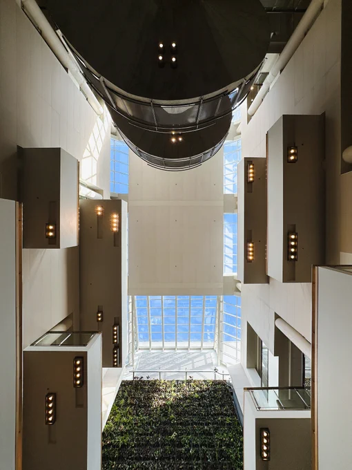 A low angle shot of a modern atrium featuring tall white walls with rectangular light fixtures and large glass windows. A lush vertical garden is visible at the base, and the ceiling is dominated by a prominent cylindrical structure. Natural light streams in, highlighting the architectural details.