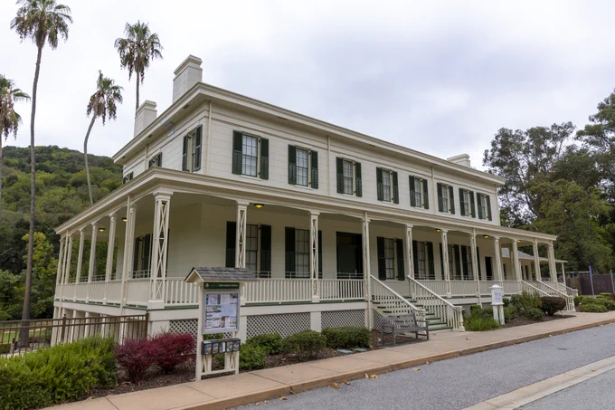 A historic two-story building with a wraparound porch and green shutters, known as Casa de Estudillo, surrounded by palm trees and landscaping. A signboard with information stands near the entrance.