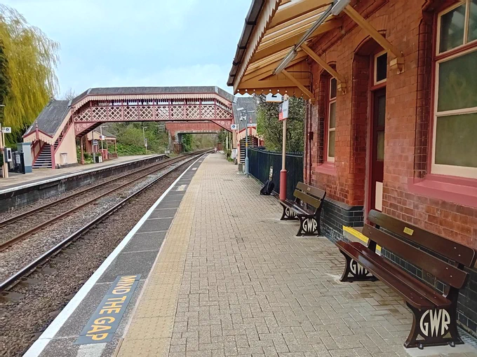 Early twentieth-century Great Western Railway station, with lattice footbridge with a cover, a roadbridge over the line in the distance, trees and greenery around, and platforms and tracks running through the centre of the image. A brick-built station building stands to the right of the image, and outside it sit two GWR-style benches.