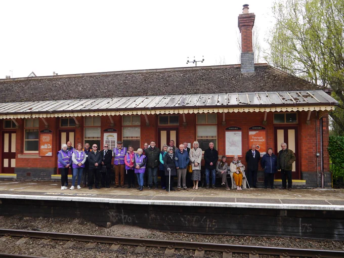 A brick-built railway station building, with canopy over the platform, stands behind a group of people. Railway tracks are to the fore.
