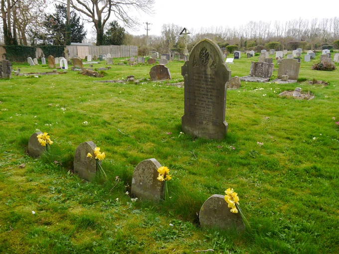 A grassed cemetery, with the image focusing on a shared headstone and four footstones. Each of the footstones has a bunch of daffodils.