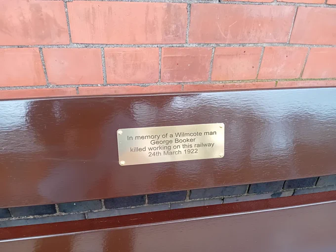 Close-up of a plaque attached to a wooden bench; it reads 'In memory of a Wilmcote man, George Booker, killed working on this railway 24th March 1922'
