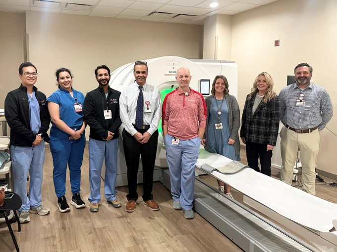 A group of medical professionals stands in a hospital room next to a PET scanner. The group consists of eight people dressed in various professional and medical attire, smiling towards the camera. The room has a neutral color scheme and modern equipment.
